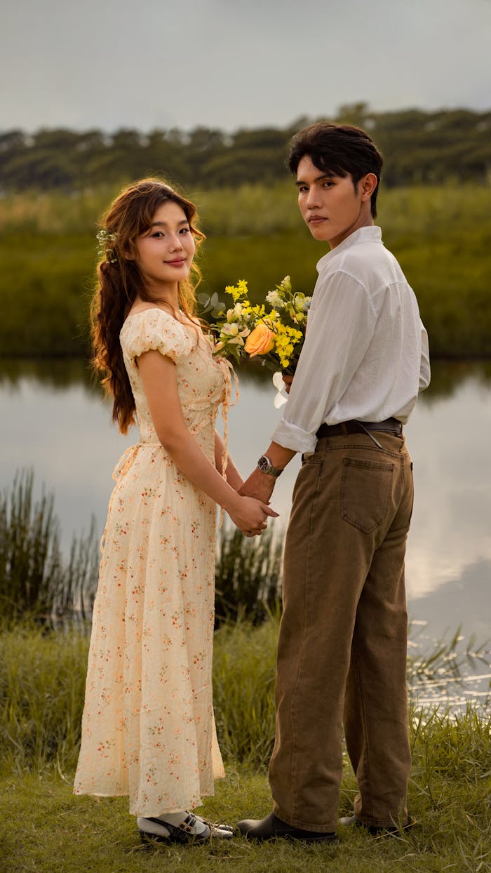 Young couple in vintage attire holding hands by a serene lakeside.